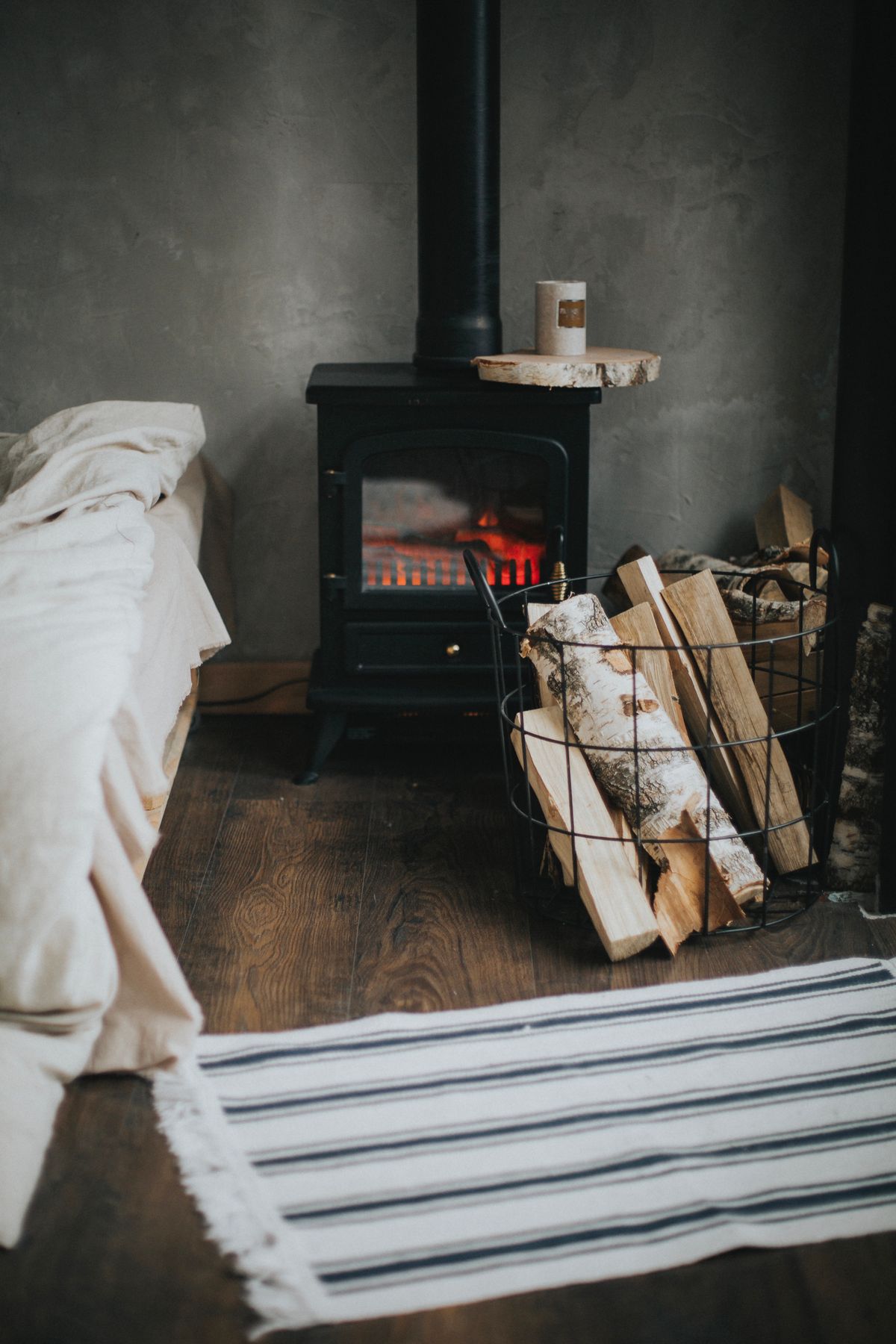 Black wood-burning stove with tall black flue pipe and glowing embers beside a wire basket of split birch firewood in a Christchurch home
