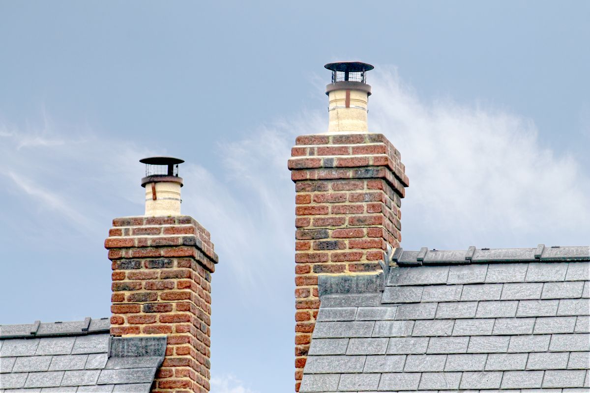 Two red brick chimney stacks with metal cowls on a grey slate roof against a pale sky